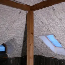 Poddasze ocieplone szarobiałą pianką natryskową, widoczne drewniane elementy konstrukcyjne i okna dachowe z niebieskim niebem w tle, zabezpieczona folią podłoga.