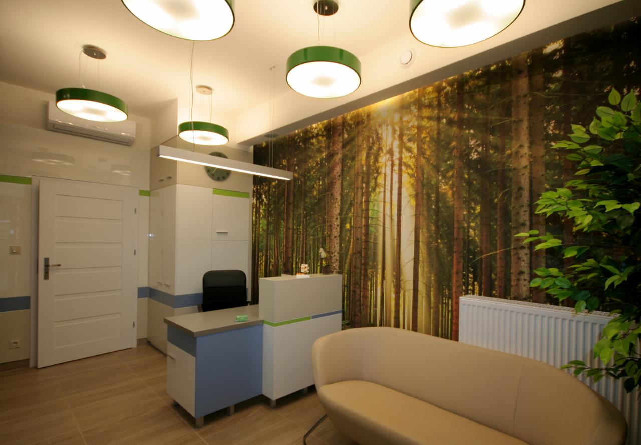 Wnętrze gabinetu medycznego z recepcją, sofą i fototapetą przedstawiającą las iglasty w ciepłych barwach, oświetlone okrągłymi lampami z zielonymi obwódkami.
