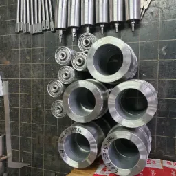 Zestaw metalowych elementów po obróbce: wałki z gwintem, tuleje, pierścienie z oznaczeniem twardości 50-55 HRC, ułożone na ciemnej powierzchni roboczej z widocznymi śladami użytkowania, obok leżą...