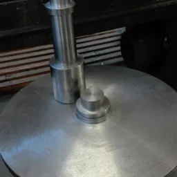 Dwa metalowe elementy obrobione na tokarce, leżące na dużym, okrągłym stole tokarskim; widoczne ślady obróbki skrawaniem.