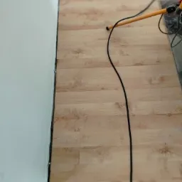 Fragment pokoju po ułożeniu jasnych paneli podłogowych, widoczna ściana z listwą przypodłogową i kabel od urządzenia na panelach. Ujęcie z góry.