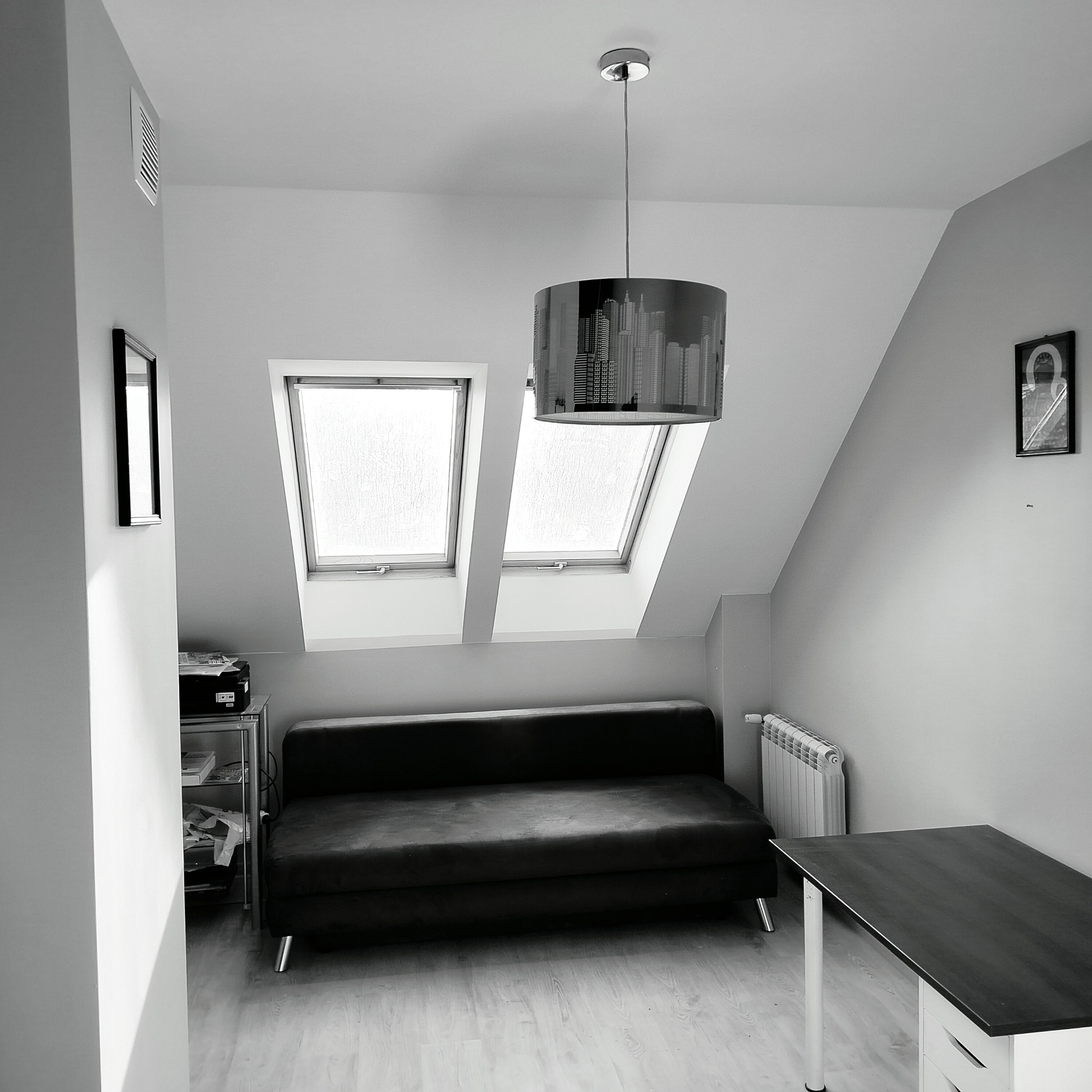 Pokój na poddaszu w szarościach: sofa, biurko, lampa z motywem miasta i dwa okna dachowe. Minimalistyczny design, stonowana kolorystyka.