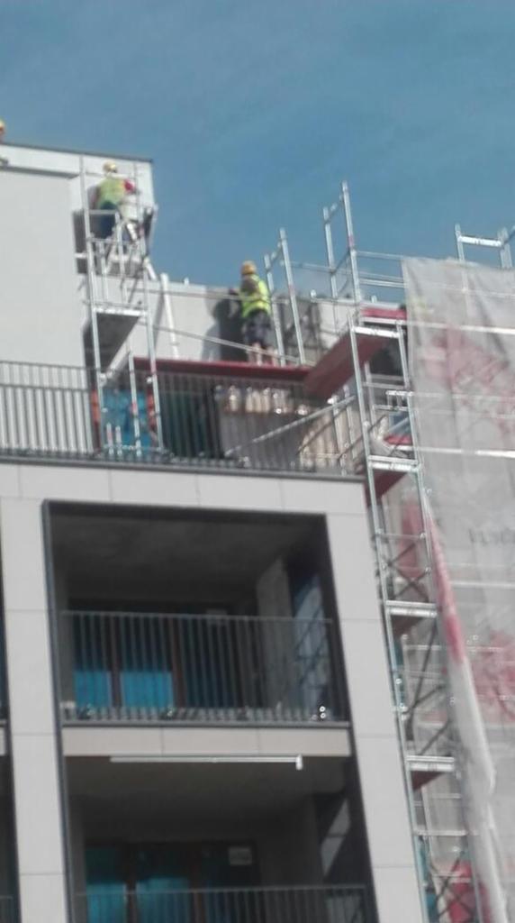 Pracownicy w żółtych kamizelkach na rusztowaniu podczas tynkowania elewacji budynku wielorodzinnego z balkonami.