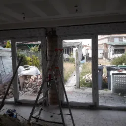 Montaż okien z białymi ramami; widoczne gruz, drabina i narzędzia w surowym wnętrzu, z widokiem na zaniedbany ogród.