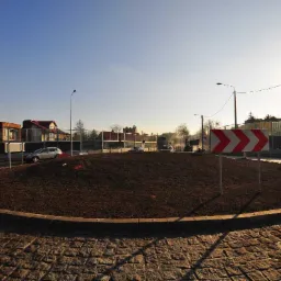 Nowo wybudowane rondo z ziemnym nasypem w centrum, otoczone kostką brukową i betonowym obrzeżem, z widocznymi znakami drogowymi i zabezpieczeniami budowlanymi w tle.