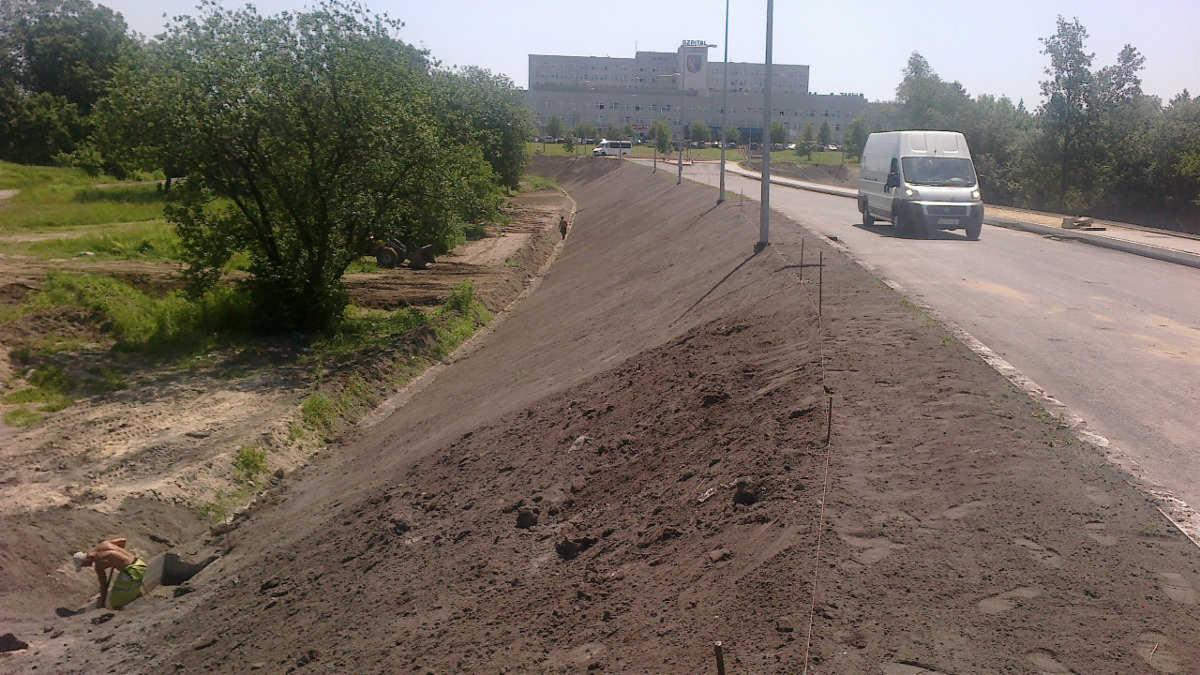 Przygotowanie gruntu pod utwardzenie drogi: wyrównany nasyp ziemny wzdłuż asfaltowej jezdni, z pracownikiem w wykopie w tle, biały van przejeżdża obok.
