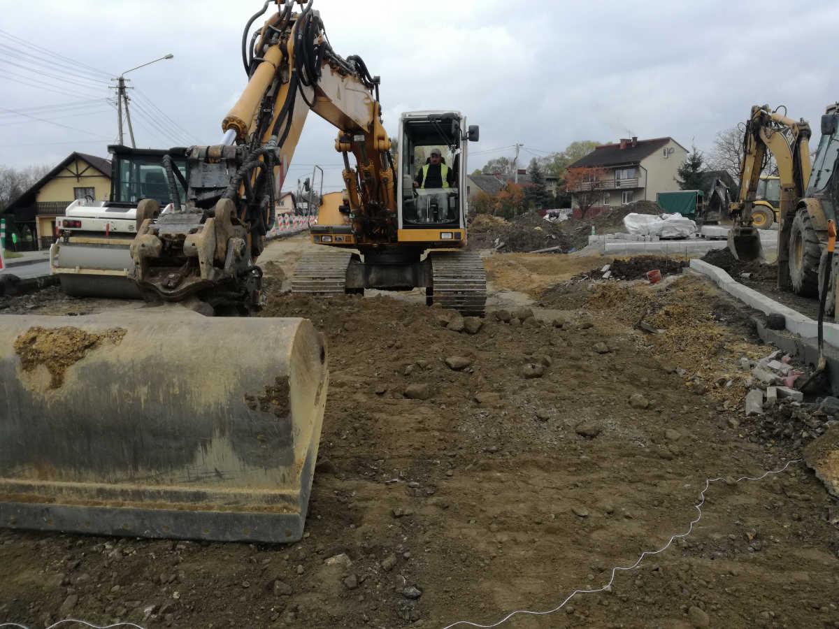 Żółta koparka z operatorem w kamizelce odblaskowej, w trakcie prac ziemnych na placu budowy, z widocznym walcem drogowym i innymi maszynami w tle, pochmurne niebo.