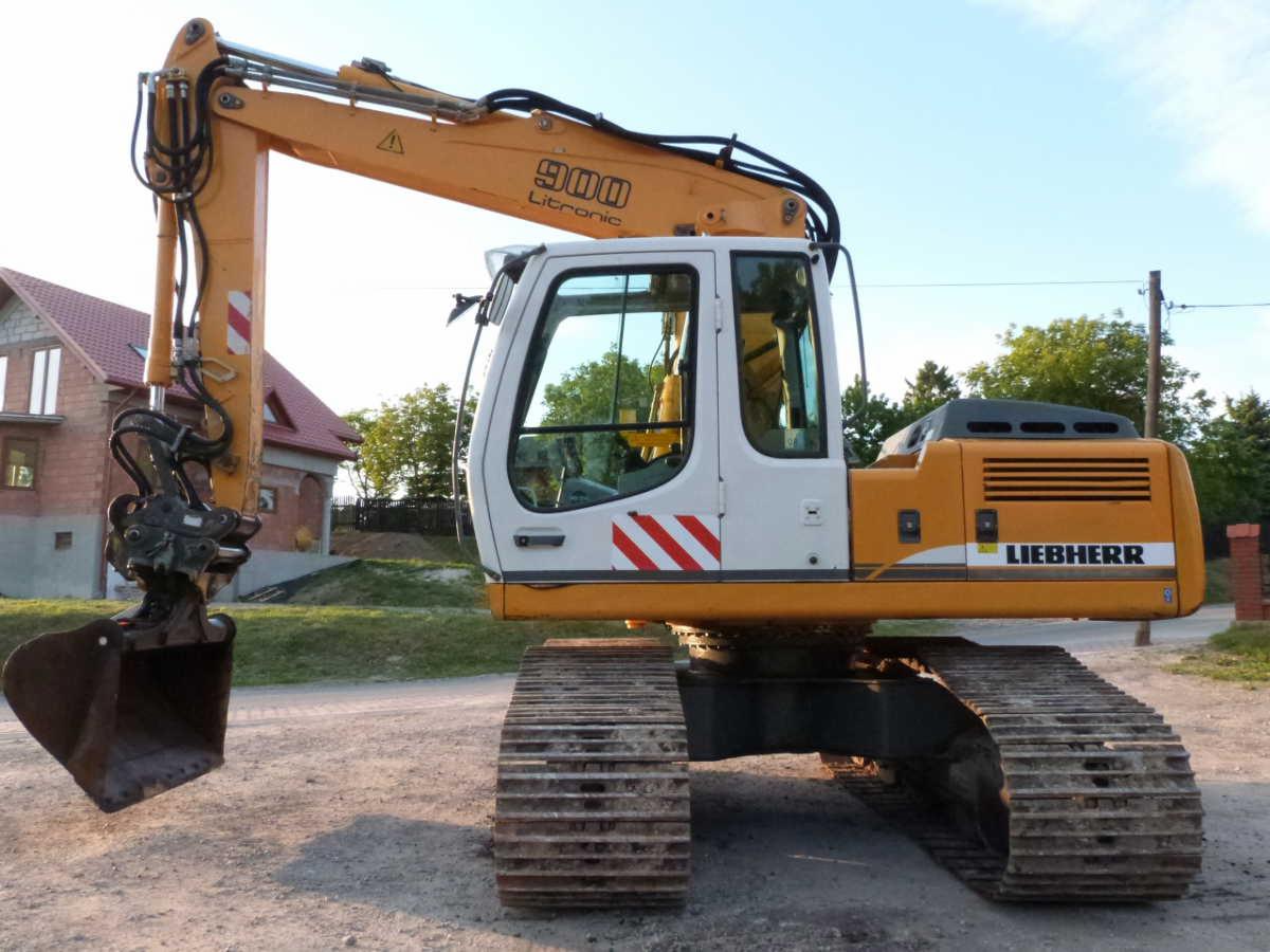 Żółto-biała koparka gąsienicowa Liebherr 900 Litronic z łyżką, stojąca na żwirowej drodze, z widocznym domem w tle.