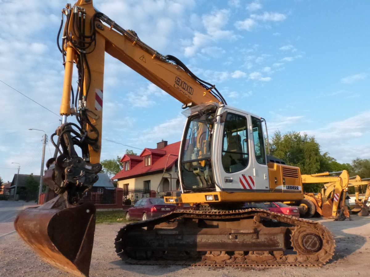 Żółto-biała koparka Liebherr 900 z łyżką, zaparkowana na żwirowym placu z widokiem na dom z czerwonym dachem w tle.