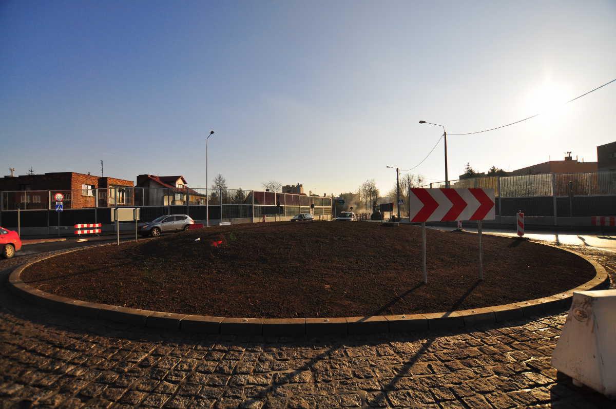 Nowo wybudowane rondo z ziemnym nasypem w centrum, otoczone kostką brukową i betonowym obrzeżem, z widocznymi znakami drogowymi i zabezpieczeniami budowlanymi w tle.