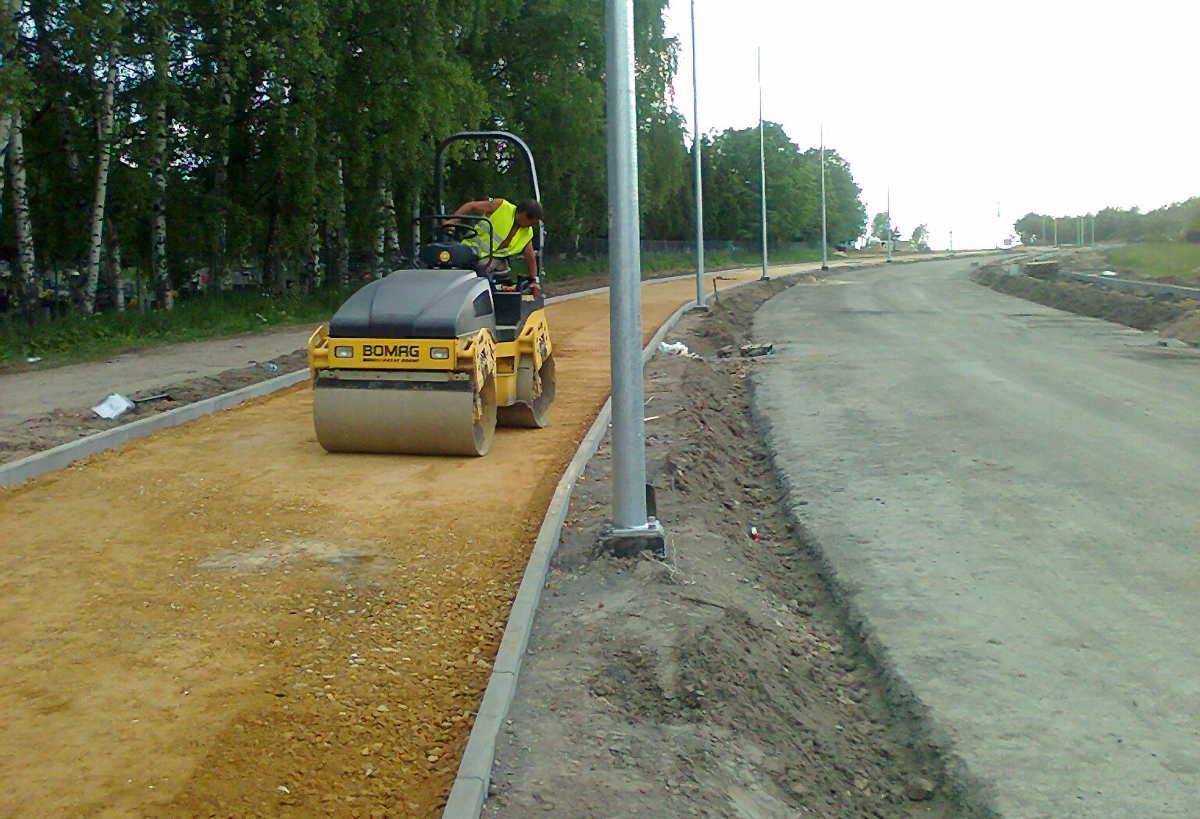 Żółty walec drogowy marki Bomag ubija piasek na nowo budowanej drodze, widoczny operator w kamizelce odblaskowej i krawężniki po obu stronach.