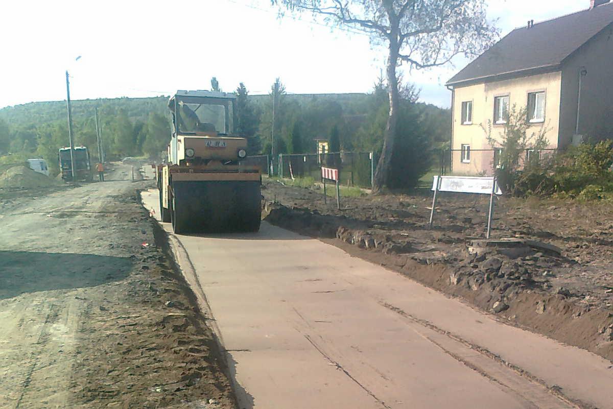 Żółty walec drogowy ubija świeżo wylaną nawierzchnię drogi gruntowej na tle zieleni i zabudowań.