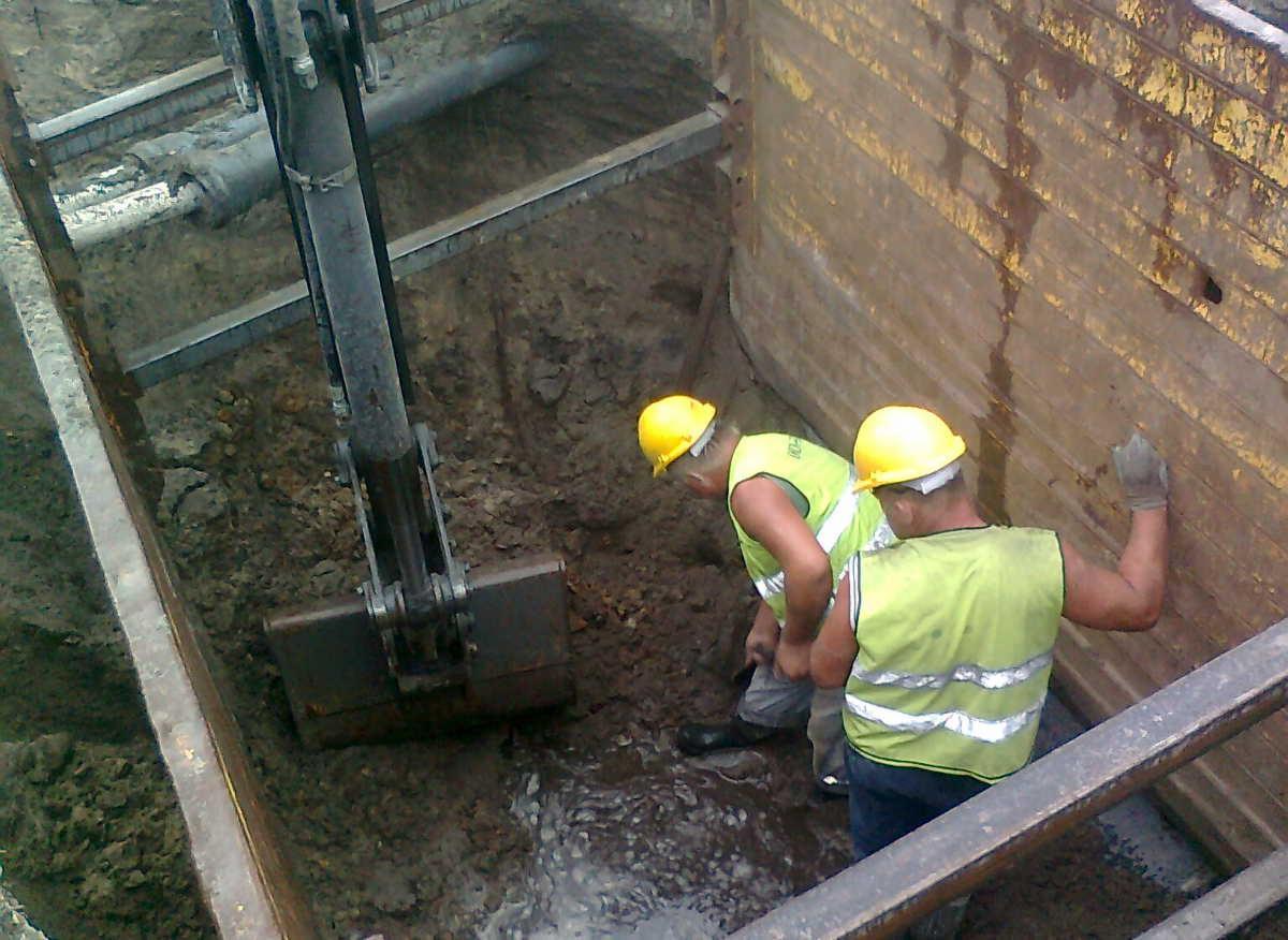 Dwóch robotników w żółtych kaskach i kamizelkach odblaskowych pracuje w głębokim wykopie zabezpieczonym metalowymi ścianami i ramami, obok stoi koparka z łyżką w błotnistej wodzie.