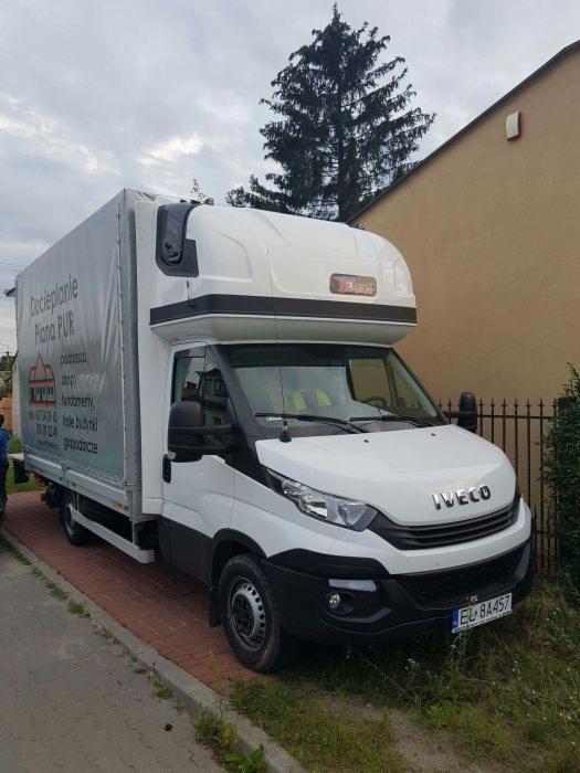 Biały samochód dostawczy Iveco z plandeką z nadrukiem reklamowym firmy świadczącej usługi docieplania pianą PUR, zaparkowany na chodniku przed budynkiem.