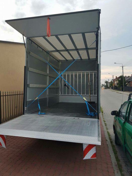 Otwarta paka busa z zabezpieczeniami ładunku w postaci niebieskich pasów i metalowej kraty, widok z zewnątrz na ulicę i zaparkowany zielony samochód.