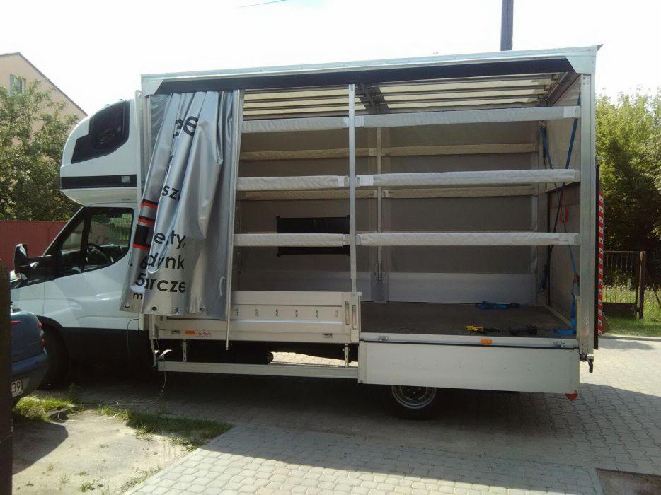 Półotwarty biały bus z półkami wewnątrz przestrzeni ładunkowej, zaparkowany na brukowanej powierzchni.