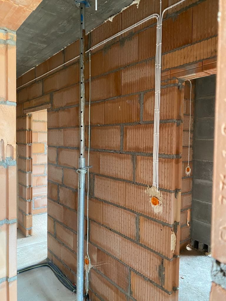 Montaż przewodów w nowym budynku z cegły. Widoczne puszki i kable prowadzone po ścianach. Szare ściany i podłoga w surowym stanie.