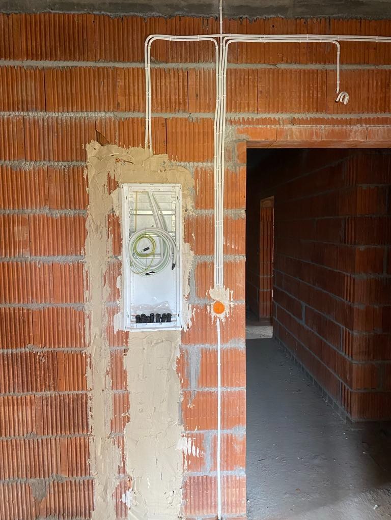 Białe przewody w bruzdach ceglanej ściany, instalacja elektryczna w puszce podtynkowej. Widoczne kable i peszle, surowy stan wykończenia wnętrza, nowa instalacja.