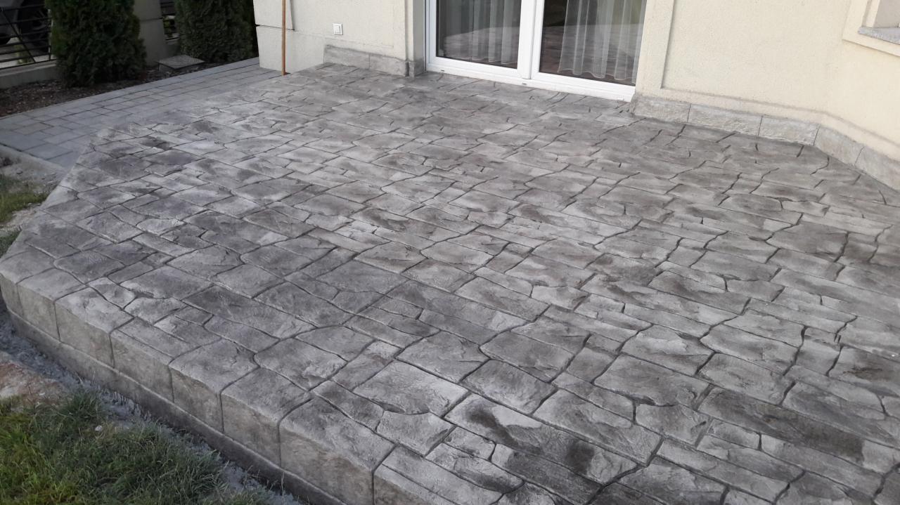 Szara, betonowa powierzchnia imitująca kamienną posadzkę, przylegająca do budynku z oknem balkonowym, z widocznymi detalami faktury i krawędzi.