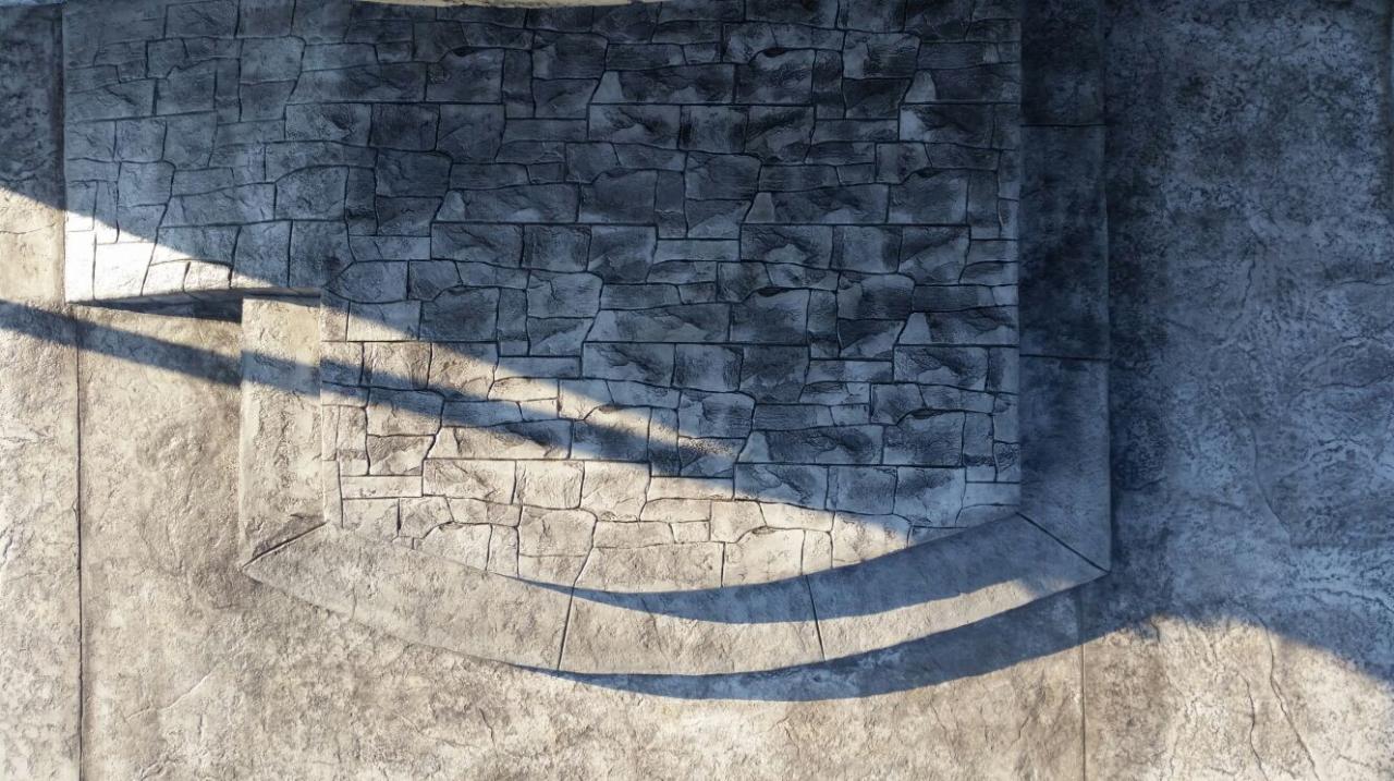 Szara ściana z betonu architektonicznego imitującego kamień, z widocznymi śladami formowania i padającym cieniem, tworząca element dekoracyjny.