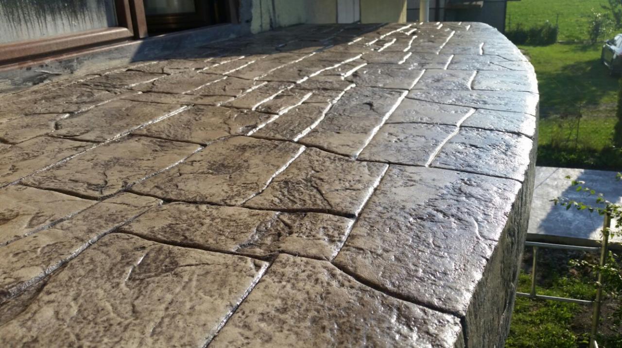 Nowa powierzchnia z betonu stemplowanego, imitująca kamienną posadzkę, przylegająca do okna budynku, z widocznym trawnikiem w tle.