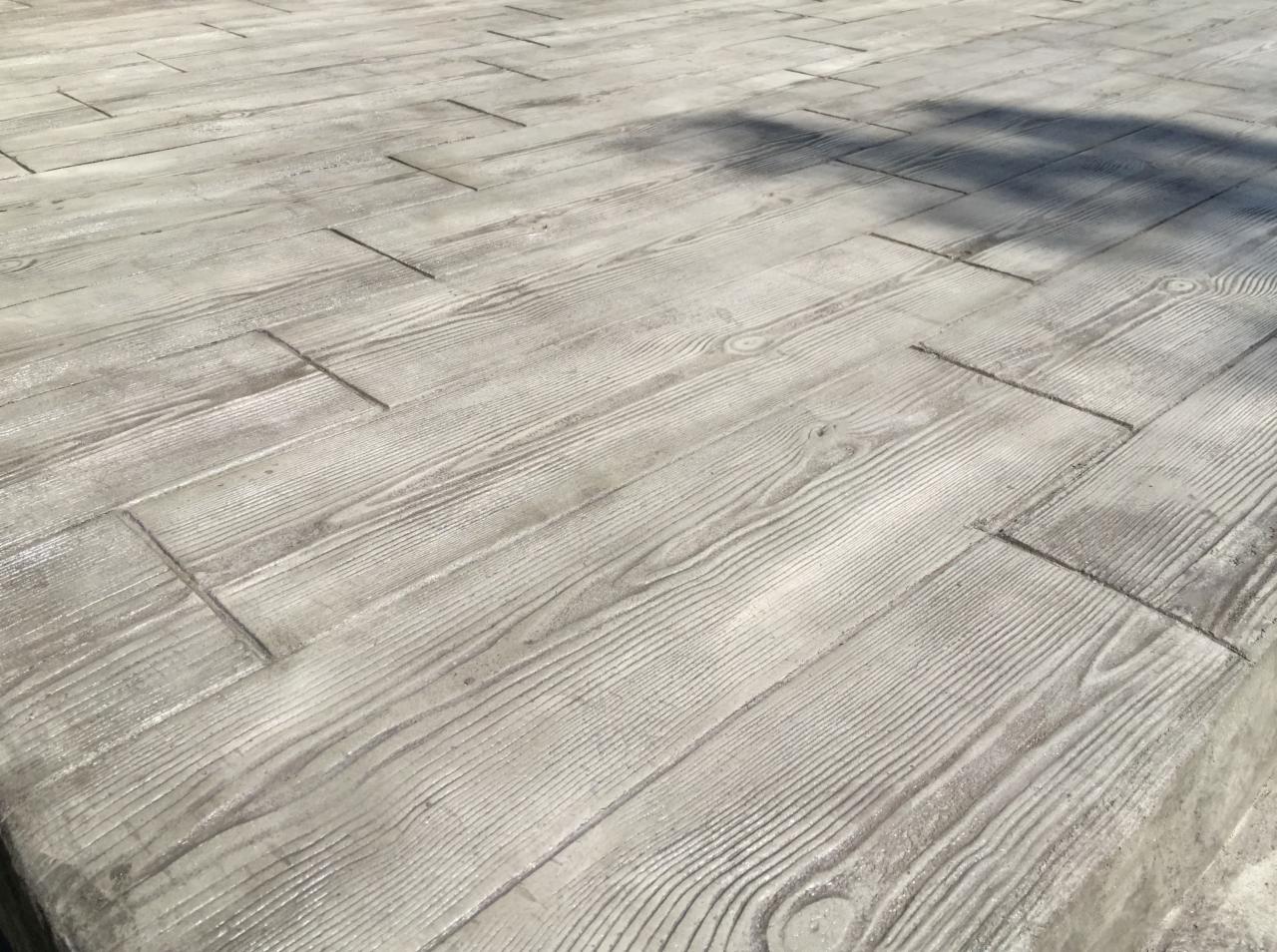 Jasnoszara, betonowa powierzchnia imitująca drewniane deski, z widocznym odciskiem słojów i zarysowanymi krawędziami, częściowo zacieniona.