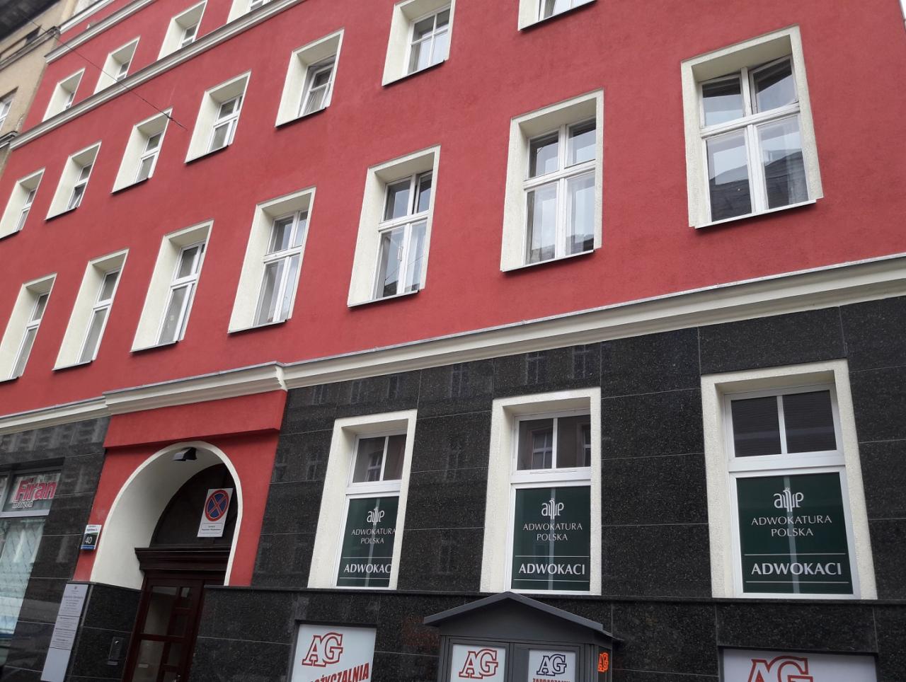 Fasada budynku z czerwonymi i czarnymi elementami, okna z naklejkami 'Adwokatura Polska Adwokaci' oraz znak zakazu parkowania nad wejściem.