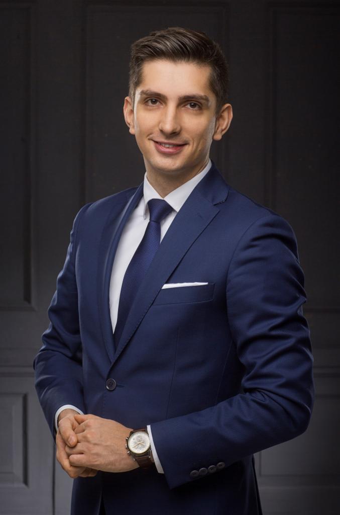 Portret uśmiechniętego mężczyzny w granatowym garniturze, białej koszuli i granatowym krawacie, na tle ciemnoszarej ściany z panelami, z zegarkiem na skórzanym pasku.