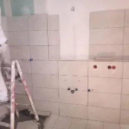 Praca glazurnicza w trakcie: pracownik na drabinie, częściowo wyłożona ściana jasnymi płytkami, widoczne instalacje wodne i otwory pod gniazdka.