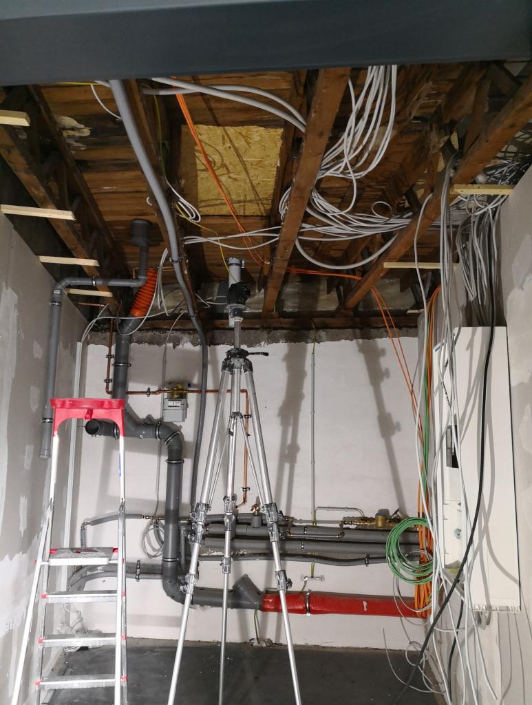 Wnętrze w trakcie remontu: widoczne instalacje elektryczne i hydrauliczne pod drewnianym stropem, drabina aluminiowa z czerwonymi elementami i statyw fotograficzny na pierwszym planie.