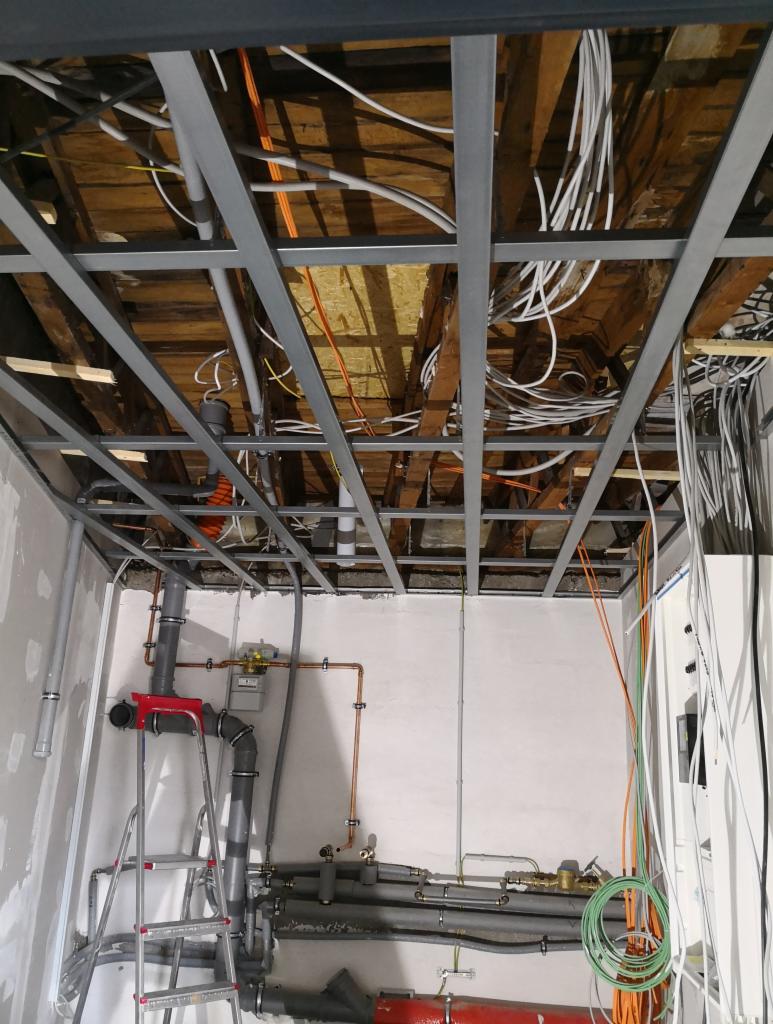 Sufit podwieszany w trakcie montażu z widocznymi instalacjami elektrycznymi i sanitarnymi, drabina oparta o ścianę, surowy stan wykończenia.