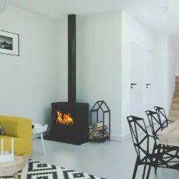 Nowoczesny salon z żółtą sofą, czarnym kominkiem wolnostojącym, geometrycznymi krzesłami i schodami w tle.