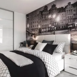 Nowoczesna sypialnia z fototapetą przedstawiającą starą architekturę miejską, białe meble z połyskiem i pościel w czarno-białe wzory geometryczne.