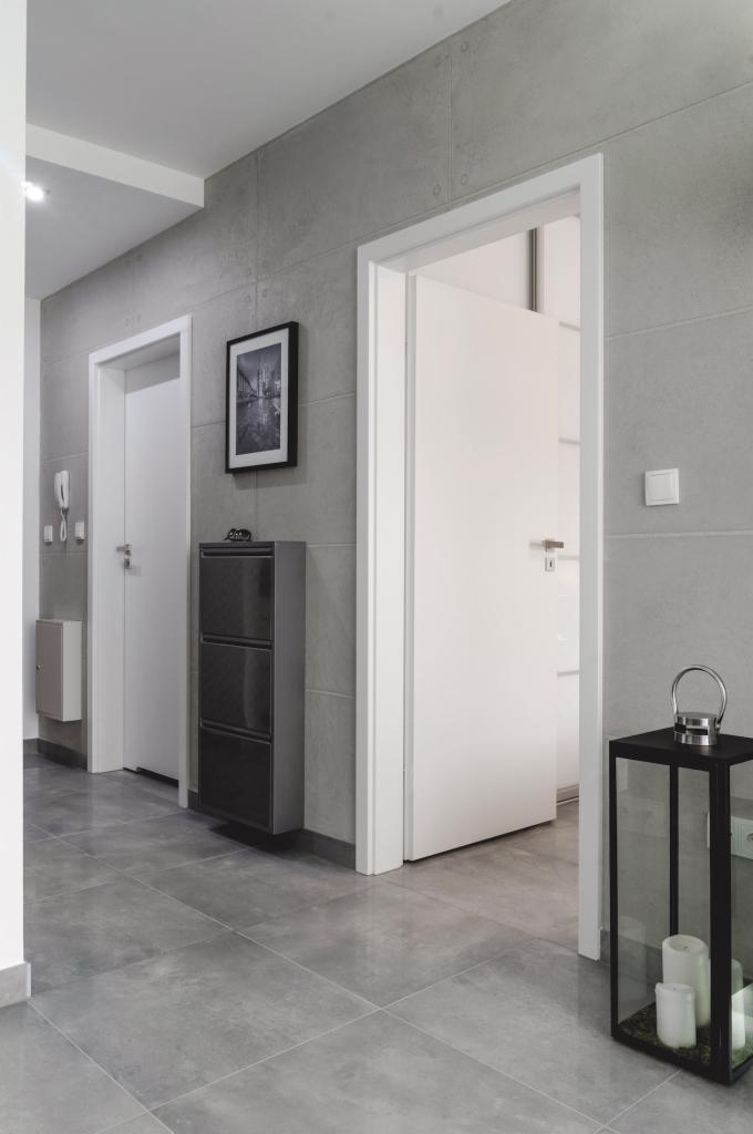 Nowoczesny przedpokój z szarą, płytkową okładziną ścian i podłogi. Widoczna szafka na buty w kolorze grafitowym, białe drzwi wewnętrzne oraz dekoracyjna latarenka z świecami na podłodze.