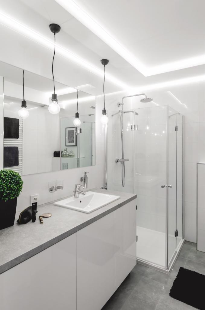 Nowoczesna łazienka w bieli i szarości. Umywalka nablatowa, kabina prysznicowa ze szklanymi drzwiami, zwisające żarówki Edison i czarny dywanik na szarej podłodze.