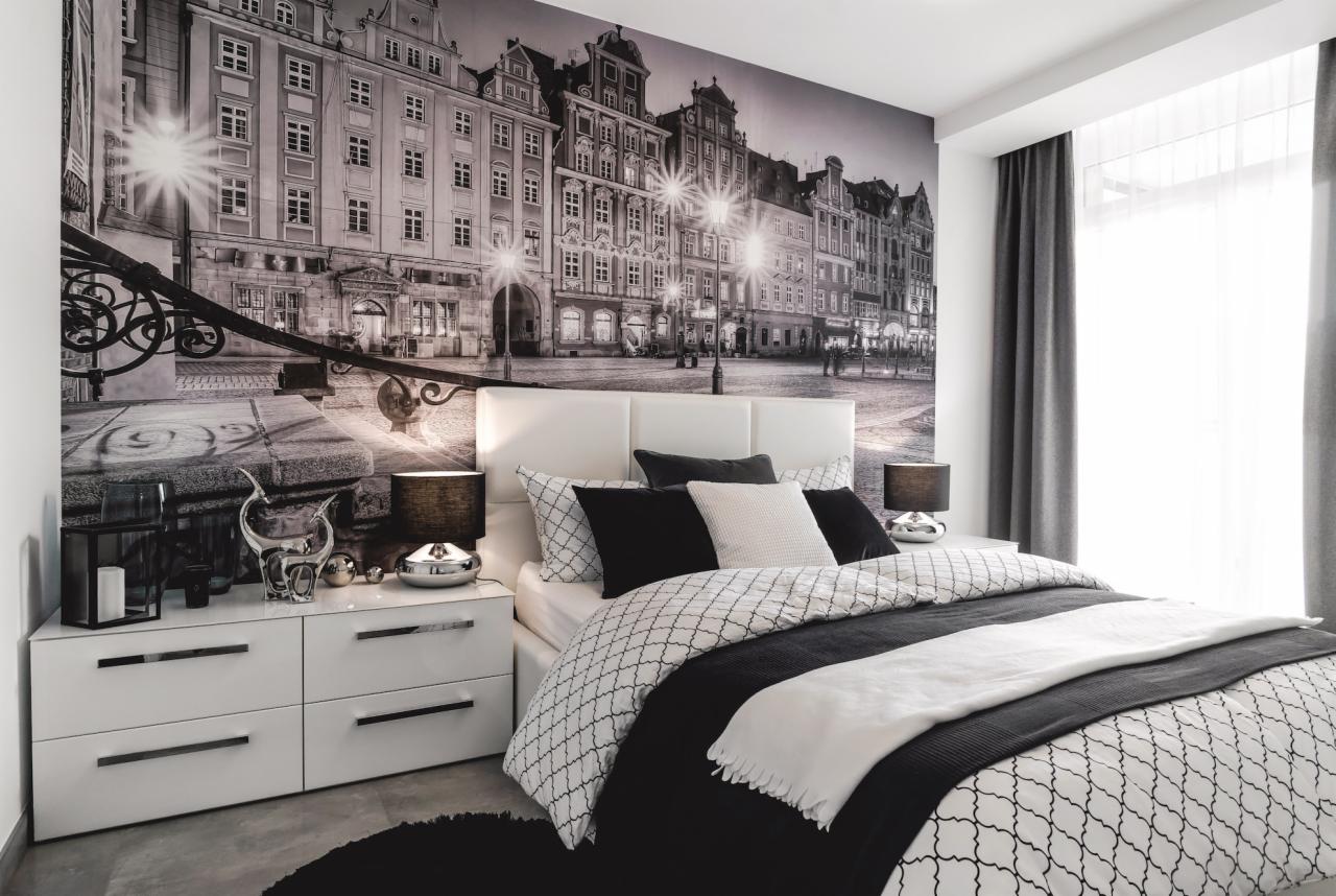 Nowoczesna sypialnia w czarno-białej tonacji z fototapetą przedstawiającą rynek miasta, biała komoda z dekoracjami i lampkami nocnymi.