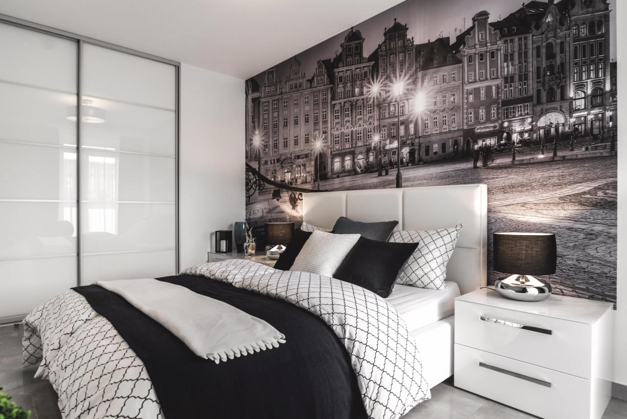 Nowoczesna sypialnia z fototapetą przedstawiającą starą architekturę miejską, białe meble z połyskiem i pościel w czarno-białe wzory geometryczne.