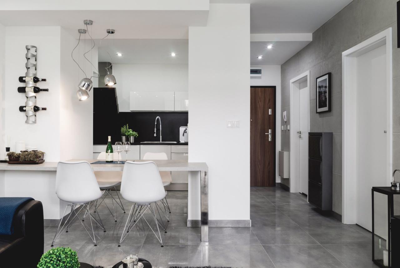 Nowoczesne wnętrze mieszkania z otwartą przestrzenią: jadalnia z białymi krzesłami Eames, minimalistyczna kuchnia z czarnym panelem, dekoracyjna półka na wino i szare płytki podłogowe.