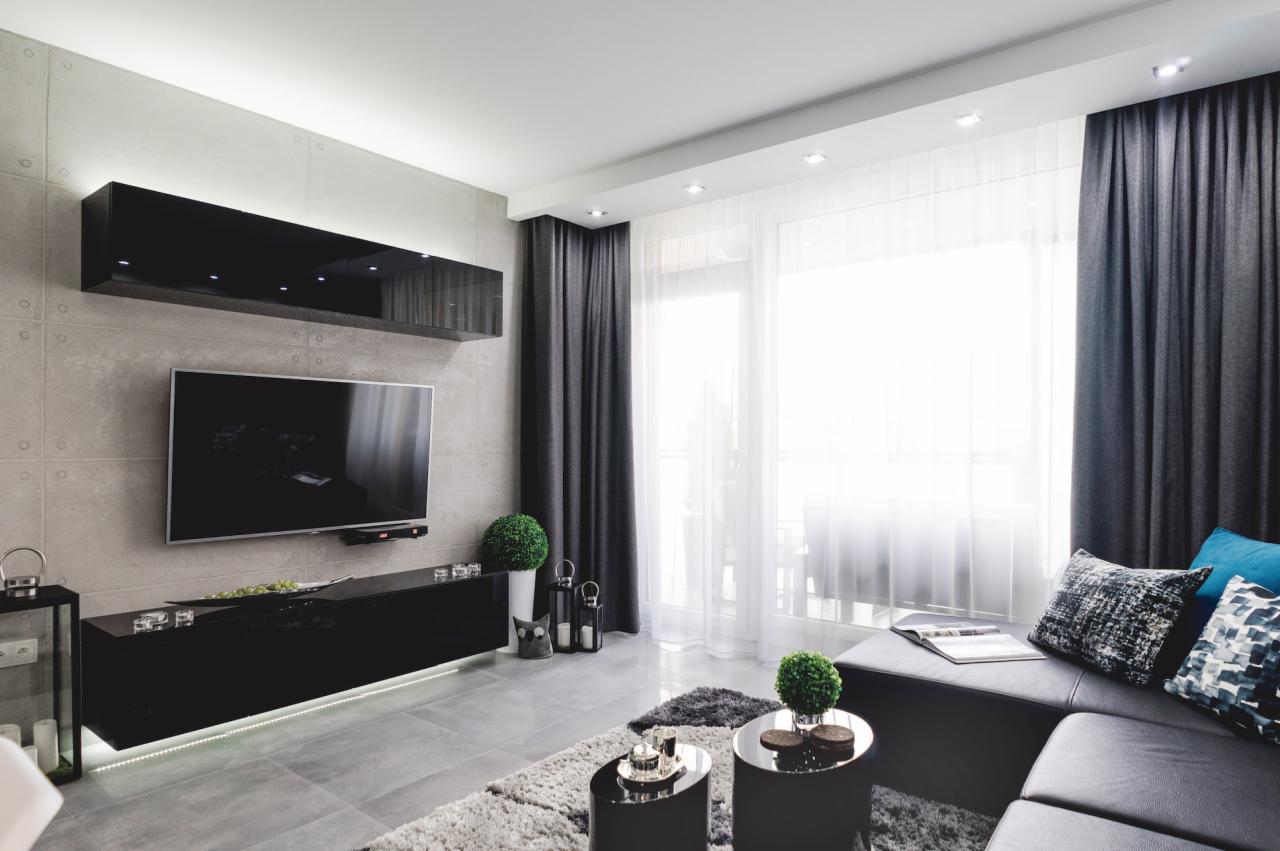 Nowoczesny salon w Gryfowie Śląskim z szarą ścianą betonową, czarnymi błyszczącymi meblami podwieszanymi z podświetleniem LED i widokiem na balkon przez okno z białymi firanami i grafitowymi...