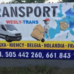 Baner reklamowy firmy transportowej Wesly-Trans z białym busem, mapą Europy z niebieskimi strzałkami i figurką pchającą wózek z kartonem, numery telefonów i kraje docelowe Polska, Niemcy, Belgia...