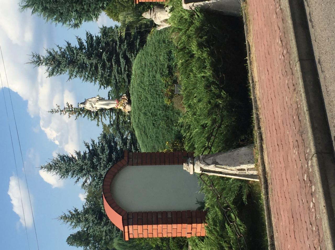 Pomnik religijny na tle zieleni i nieba, widoczny fragment muru z czerwonej cegły