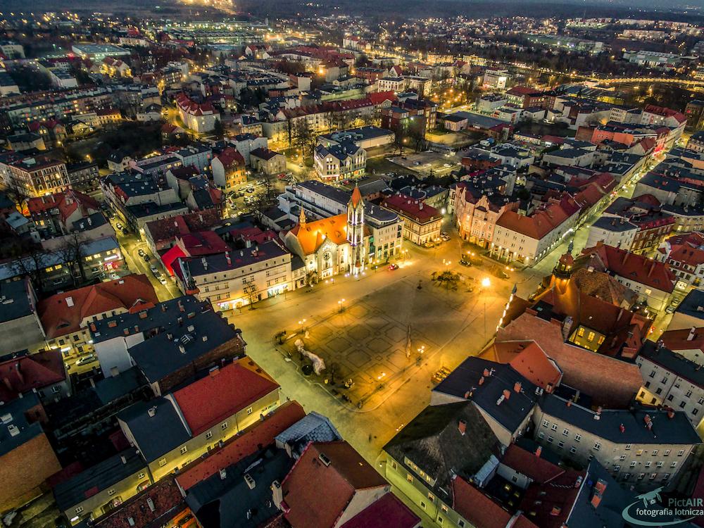 Panorama miasta nocą z lotu ptaka, oświetlony rynek z kościołem w centrum, czerwone dachy budynków, światła uliczne.