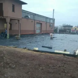 Przygotowanie podłoża pod budynek: rozłożona czarna folia izolacyjna na warstwie styropianu, widoczni pracownicy i niwelator laserowy.