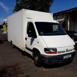 Biały samoch&oacute;d dostawczy Renault Master z zabudową kontenerową zaparkowany na asfalcie, widoczny numer rejestracyjny DLE-06872, obok budynek magazynowy i zaparkowany samoch&oacute;d osobowy, słoneczny dzień.