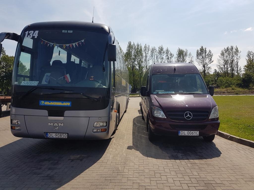 Szary autokar MAN z dekoracjami z flag i bordowy bus Mercedes Sprinter zaparkowane obok siebie na brukowanym placu w słoneczny dzień.