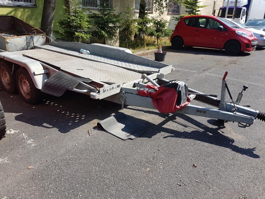 Dwupoziomowa, stalowa przyczepa samochodowa z najazdami, zaparkowana na asfaltowym placu, z widocznym czerwonym samochodem w tle.