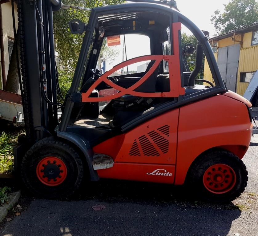 Czerwono-szary wózek widłowy Linde stoi na asfalcie przed żółtym budynkiem gospodarczym, widoczne logo firmy na boku.