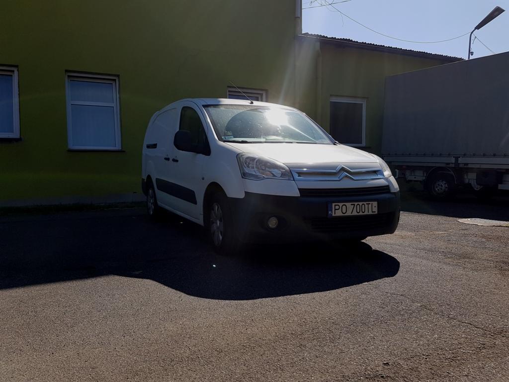 Biały samochód dostawczy marki Citroen Berlingo zaparkowany na asfaltowym placu przed budynkiem o oliwkowym kolorze, z widoczną tablicą rejestracyjną PO 700TL i odblaskami słońca na przedniej szybie.