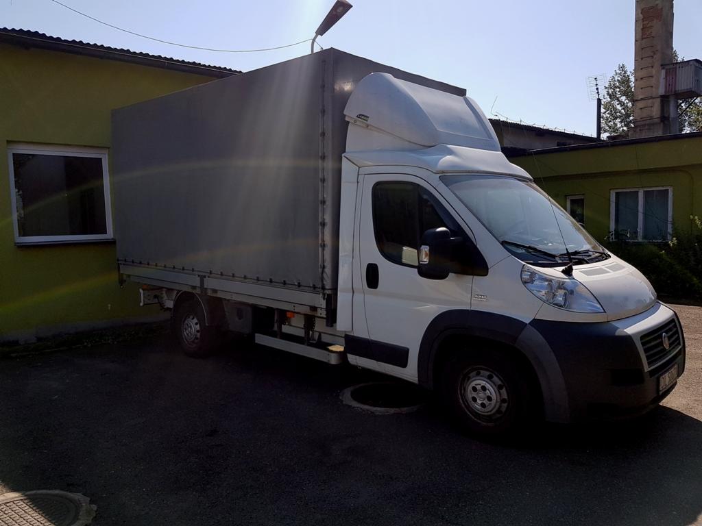 Biały samochód dostawczy z plandeką na tle zielonego budynku z oknem. Widoczny komin w tle.