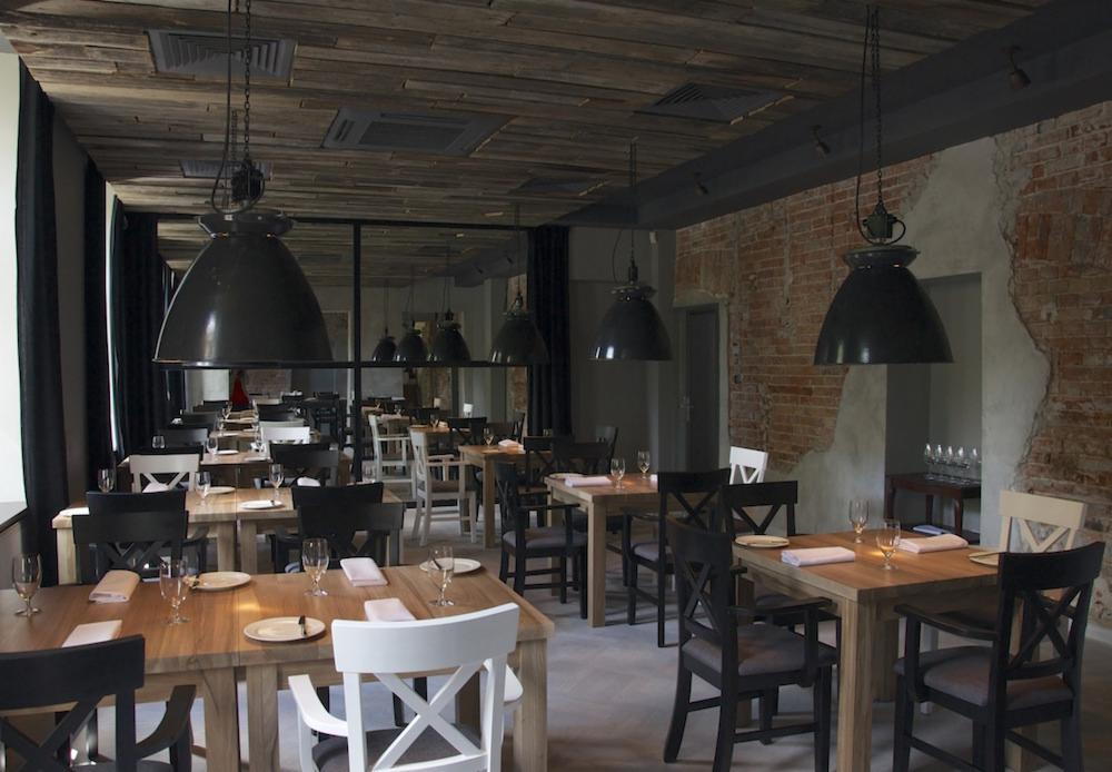 Wnętrze restauracji z surowymi ścianami z cegły, drewnianym sufitem i dużymi, wiszącymi lampami w stylu industrialnym nad nakrytymi stołami.